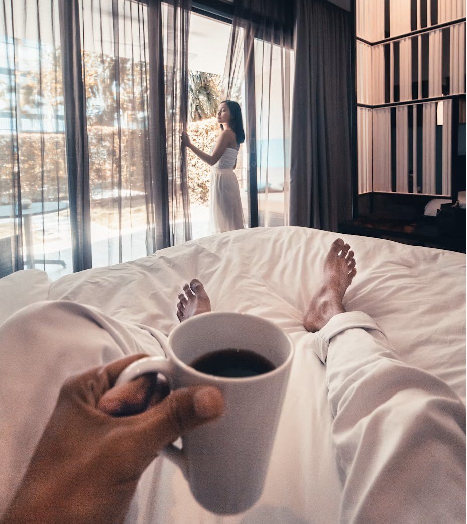 Pies descalzos sobre la cama sujetando un café, mientras alguien abre las cortinas al fondo