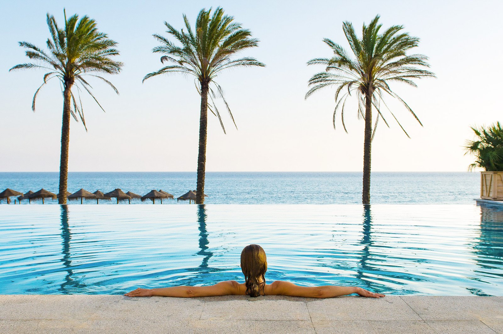 Huésped sumergida en una piscina infinita frente al mar, entre tres palmeras, al atardecer